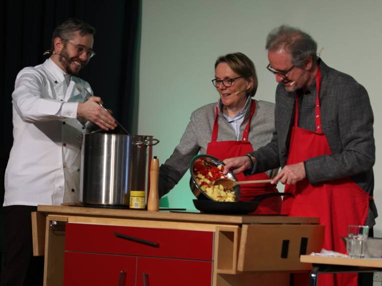 Drei Personen - zwei Männer und eine Frau - tragen rote Schürzen und stehen hinter einer mobilen Kochstelle. 