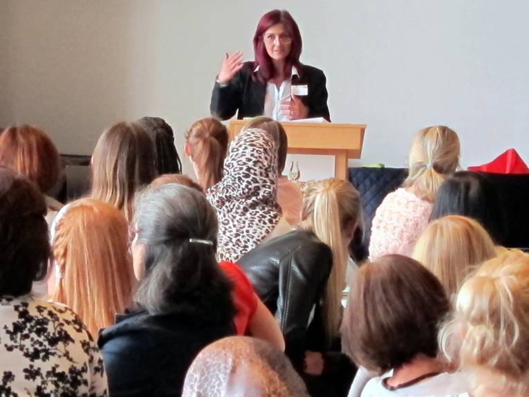 Eine Frau steht hinter einem Rednerpult und spricht. Vor ihr sitzen Zuschauer/innen und hören ihr zu.