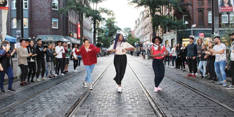Etwa 20 Jugendliche stehen in zweit Linien am Rand einer Straße. In der Mitte stehen drei Mädchen nebeneinander und überkreuzen die Füße, um einen Seitwärtsschritt zu machen.