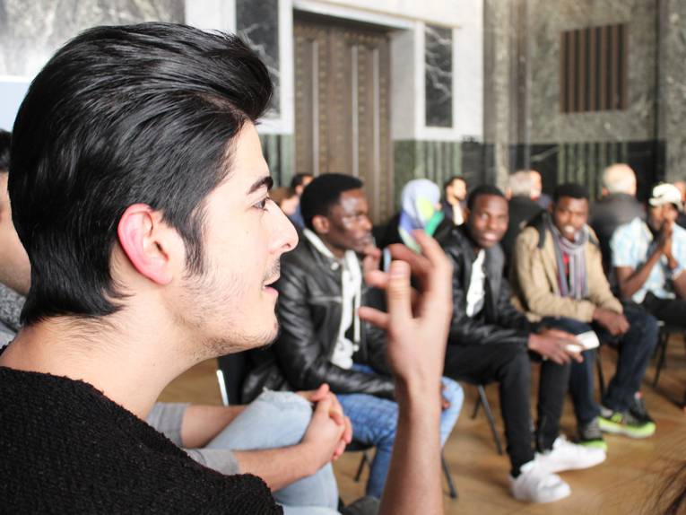 Ein Mann sitzt im Hodlersaal und spricht, während er mit seiner rechten Hand gestikuliert. Hinter ihm sind etwa zwölf weitere Personen erkennbar, die in seinem, oder dem benachbarten Stuhlkreis sitzen.