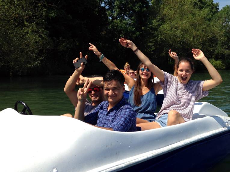 Vier Jugendliche sitzen in einem Tretboot und winken in die Kamera. Dahinter sind zwei Personen auf einem weiteren Tretboot zu erkennen.