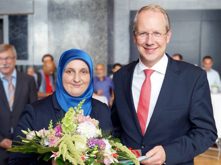 Zwei Personen stehen nebeneinander und lächeln. Die Frau (links im Bild) trägt einen Blumenstraß in der Hand.