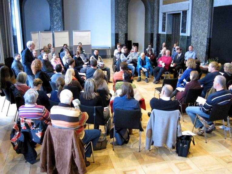 Etwa 50 Personen sitzen Kreisförmig im Mosaiksaal. Eine Person steht und spricht.