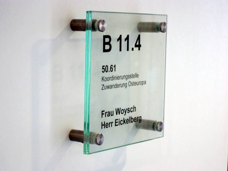 Foto eines transparenten Büroschildes, auf dem steht "Koordinierungsstelle Zuwanderung Osteuropa und die Namen "Frau Woysch" und "Herr Eickelberg".