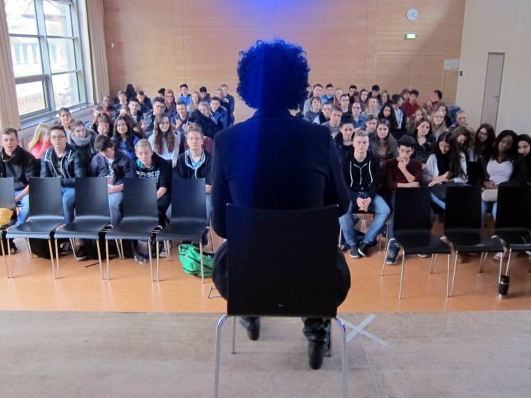 Eine Person sitzt auf einer Bühne und blickt etwa 70 Schüler/innen entgegen.