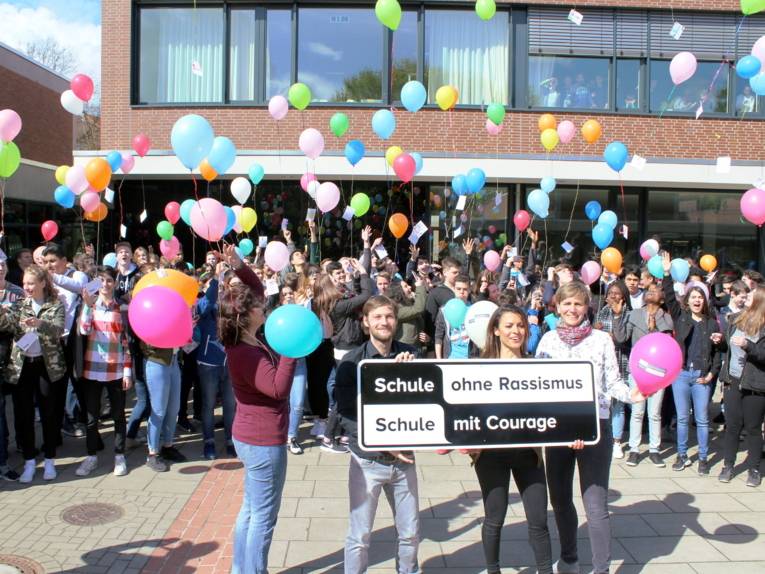 Etwa 80 Schüler/innen stehen hinter vier Lehrer/innen die das typische SOR-SMC-Banner vor sich halten. Die Schüler/innen lassen Ballons in den Himmel steigen, an denen Postkarten befestigt sind.