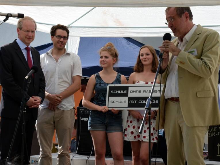 Fünf Personen stehen auf einer Bühne im Freien nebeneinander. Die rechte Person spricht in ein Mikrofon und schaut in Richtung der ganz links stehenden Person.