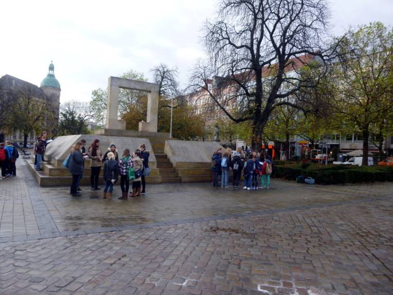 Etwa 20 SchülerInnen stehen verteilt in drei Kleingruppen um das Mahnmal am Opernplatz verteilt und hören den jeweiligen Referent/innen zu.