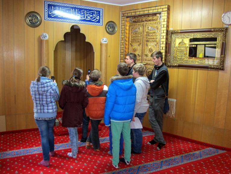 Zwei ältere Schüler erklären fünf jüngeren Schüler/innen die Mihrab. Zwei der Zuhörenden halten ihre Handys hoch, um zu fotografieren.