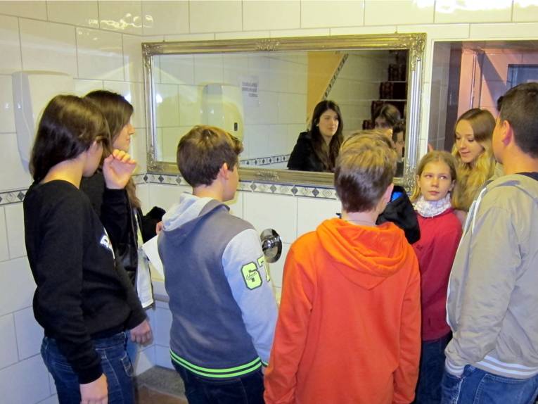 Sieben Jugendliche stehen vor zwei Waschbecken nebeneinander und einem breiten Spiegel darüber. Rechts im Bild spricht eine Schülerin und informiert die Jüngeren.