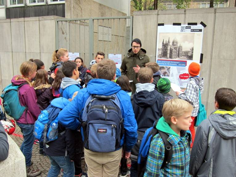 Ein Mann steht vor einer Gruppe von Schüler/innen neben einem Infoplakat, auf dem das SOR-SMC-Banner und eine Zeichnung der zerstörten Synagoge erkennbar ist. 
