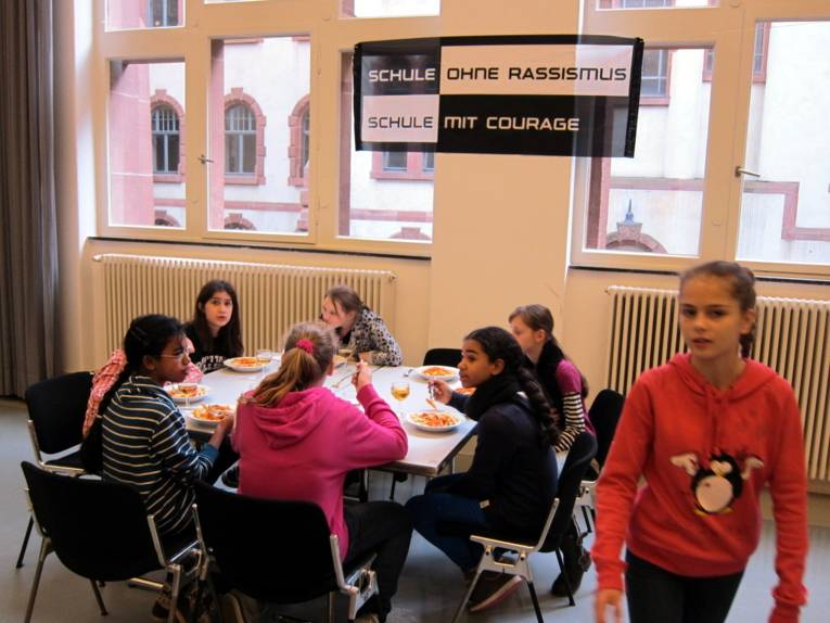 Sieben Schüler/innen sitzen um einen Tisch im Bürgersaal und essen. Über ihnen hängt das SOR-SMC-Banner. Ein Mädchen steht und läuft in Richtung Ausgang.