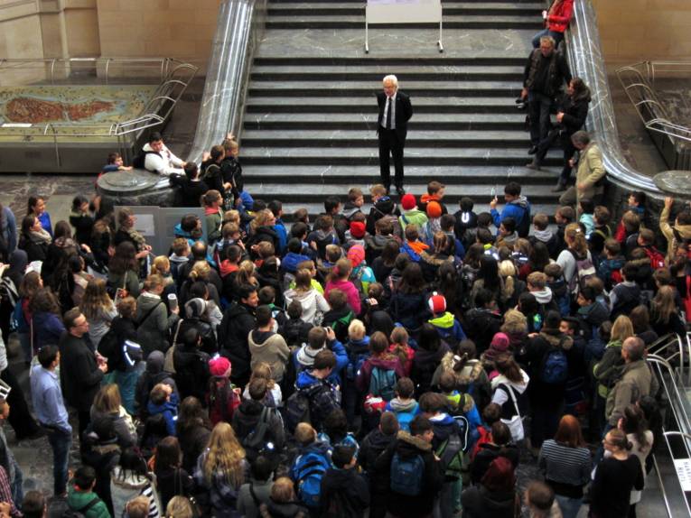 Der Bürgermeister steht auf der Rathaustreppe und spricht zu den Schüler/innen. Diese bilden einen Menschentraube unter der Treppe. Einige recken die Hälse, um besser zu sehen. Oben rechts am Rand neben Strauch auf der Treppe stehen drei bis vier Journalisten.
