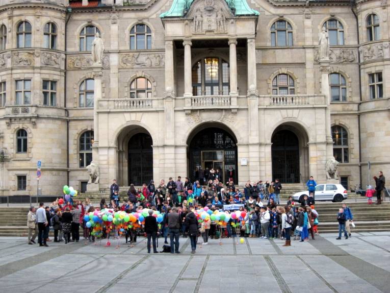 Veranstalter/innen und Schüler/innen sammeln sich vor dem Rathaus. Vor der Menschenmenge sind mit kleinen Gewichten befestigte Ballons, die gleich verteilt werden.