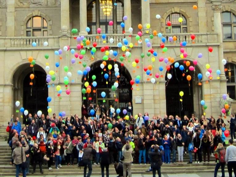 Schüler/innen und Leher/innen stehen vor dem Neuen Rathaus und lassen die etwa 300 Ballons mit den Postkarten steigen. Die meisten schauen nach oben. Hinter den Ballons ist die Rathaus-Fassade zu erkennen.