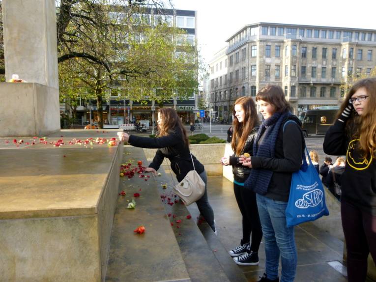 Vier Mädchen stehen in einer Reihe vor dem Denkmal und eine legt Blumenblüten aufs Denkmal. Eine andere hält Blüten ind er Hand.