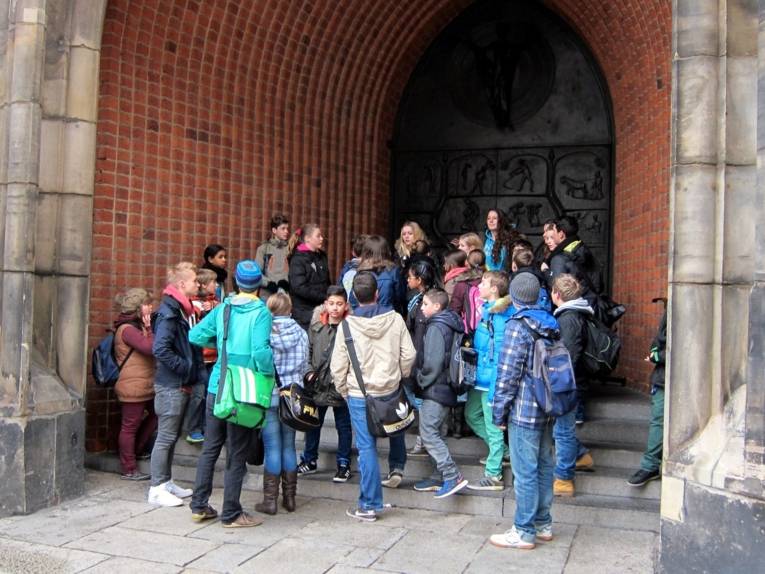 Etwa 20 Schüler/innen stehen am Eingangstor der Marktkirche und lassen sich von anderen Schüler/innen ihre Architektur und Verzierung erklären.