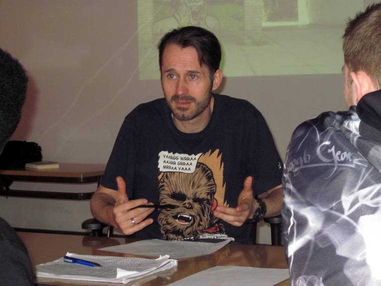 Ein Mann mit T-Shirt sitzt Schüler/innen gegenüber und gestikuliert mit beiden Händen beim Sprechen.
