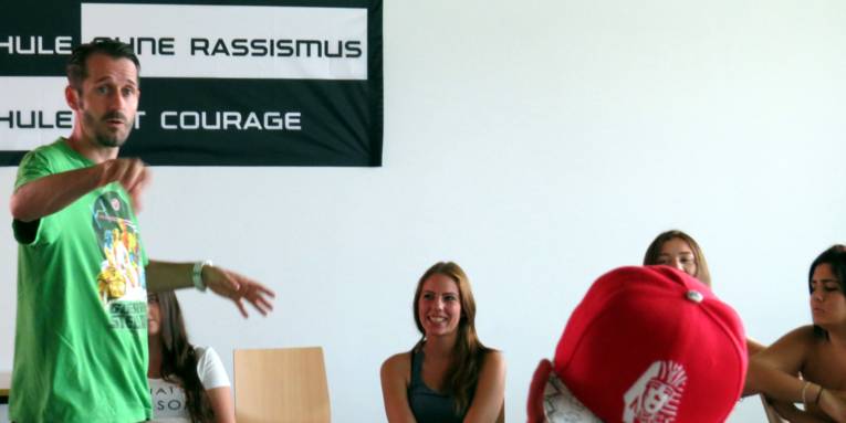 Spax steht vor dem SOR-SMC-Banner und spricht, während er mit dem Zeigefinger in Richtung der Kamera zeigt. 5 Schüler/innen sitzen im Kreis und hören ihm zu.