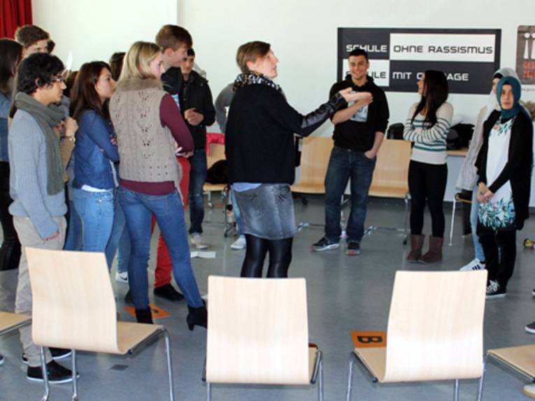 Etwa 15 Schüler/innen stehen. Eine steht in der Mitte und spricht. Die anderen schauen sie an.