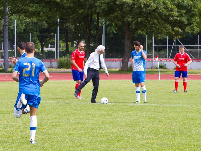 Ein Mann in Hemd und Anzughose führt einen Anstoß beim Fußball aus, um ihn herum Spieler in roten und blauen Trikots