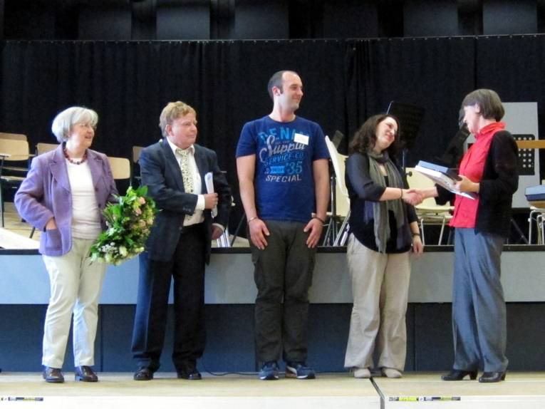 Vier Personen stehen nebeneinander auf einer Bühne. Die linke Frau hält einen Blumenstraß in der Hand, während die Rechte Frau der zweiten Frau von rechts die Hand schüttelt. 