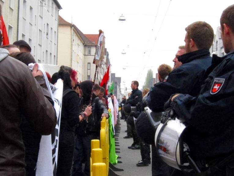 Auf der Linken Seite sind Aktivist/innen, die durch eine gelbe Kunststoffsperren von der nördlichen Straßenseite und den Gleisen fergehalten werden. Auf der rechten Seite der Absperrungen stehen Polizist/innen.