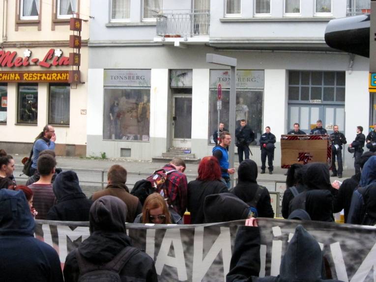 Im Vordergrund stehen einige Demonstrierende mit Fahnen und Transparenten. Vor ihnen ist das Thor-Steinar-Geschäft, das zuvor mit Farbbeuteln und Eiern beworfen wurde.