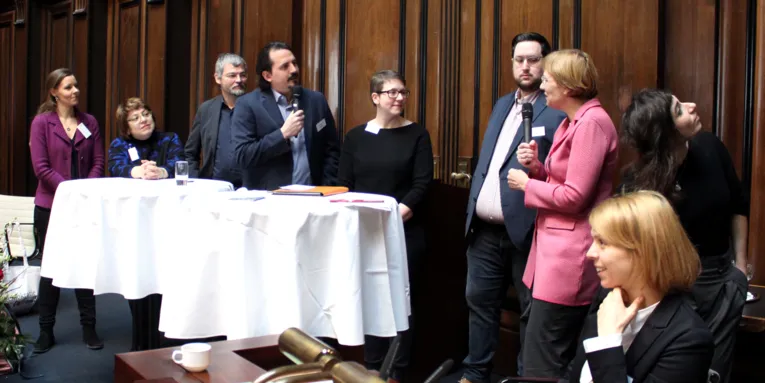 Acht Personen - fünf Frauen und drei Männer - stehen im Hodlersaal des Neuen Rathauses hinter Stehtischen. Ein Mann und eine Frau halten Mikrofone in der Hand und blicken zueinander. Rechts daneben sitzt eine weitere Frau.