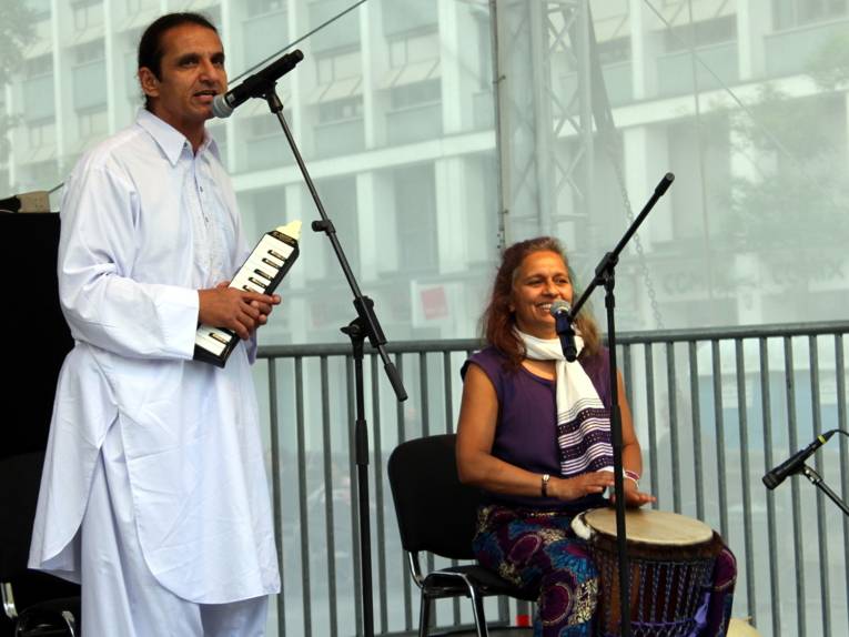 Ein Mann und eine Frau stehen und sitzen auf der Bühne und spielen auf Instrumenten. Der stehende Mann trägt einen traditionellen afghanischen Pirhan Tumban und singt.