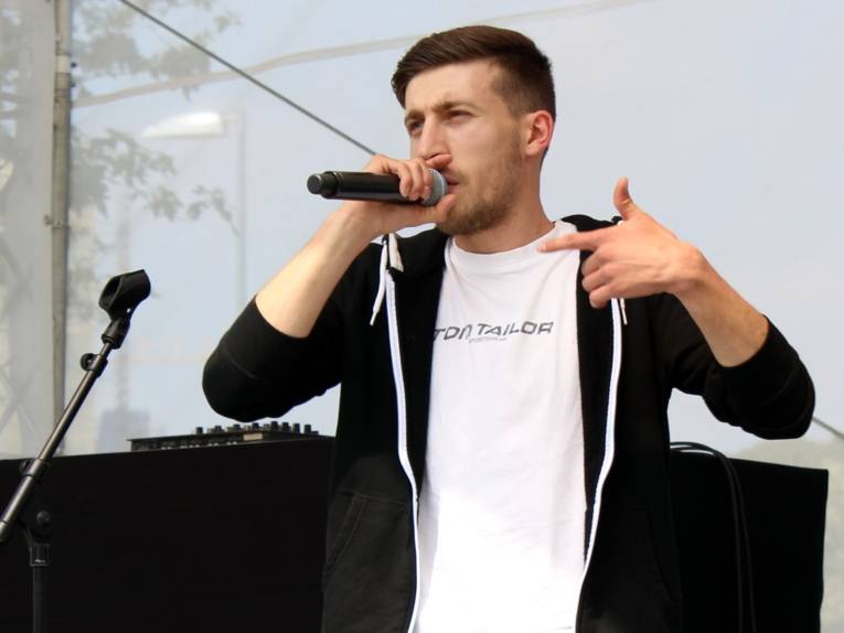 Ein Mann steht auf einer Bühne und singt in ein Mikrofon, das er dicht vor dem Mund hält. Mit dem Zeigefinger seiner anderen Hand zeigt er nach vorn.