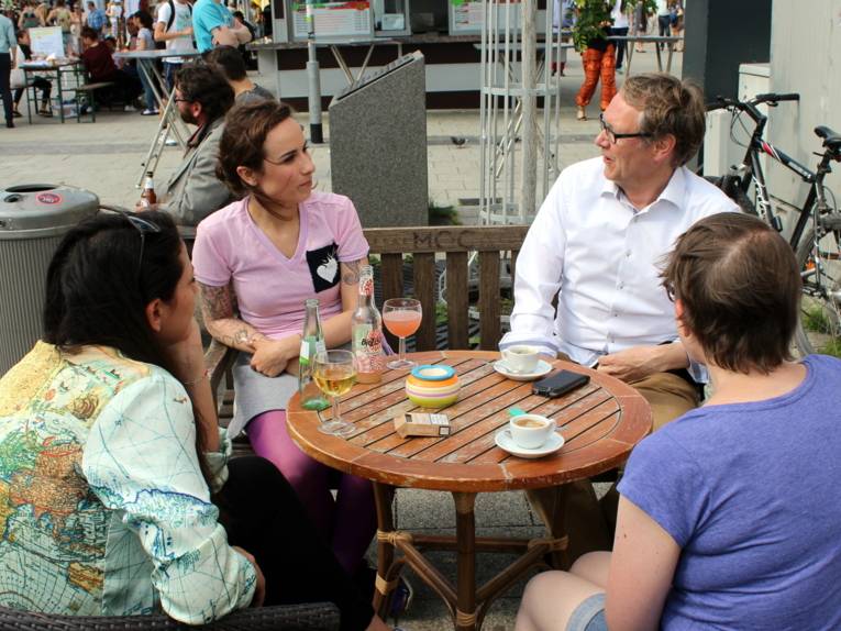 Vier Personen - drei Frauen und ein Mann - sitzen in am nördlichen Ende des Goseriedeplatzes am Tisch eines Cafés und unterhalten sich. Auf dem Tischen stehen Kalt- und Heißgetränke.