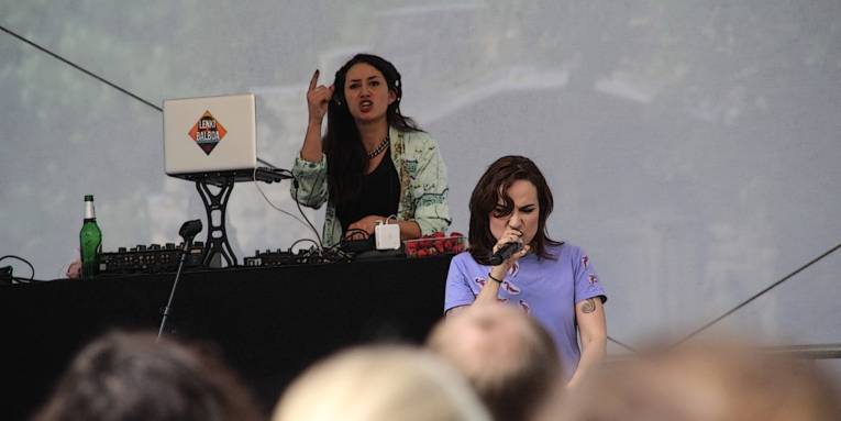 Zwei Frauen stehen auf der Bühne. Eine davon steht hinter einem aufgeklappten Notebook, blickt nach vorne und hält einen Zeigefinger hoch. Die andere steht rechts daneben und singt in ein Mikrofon.