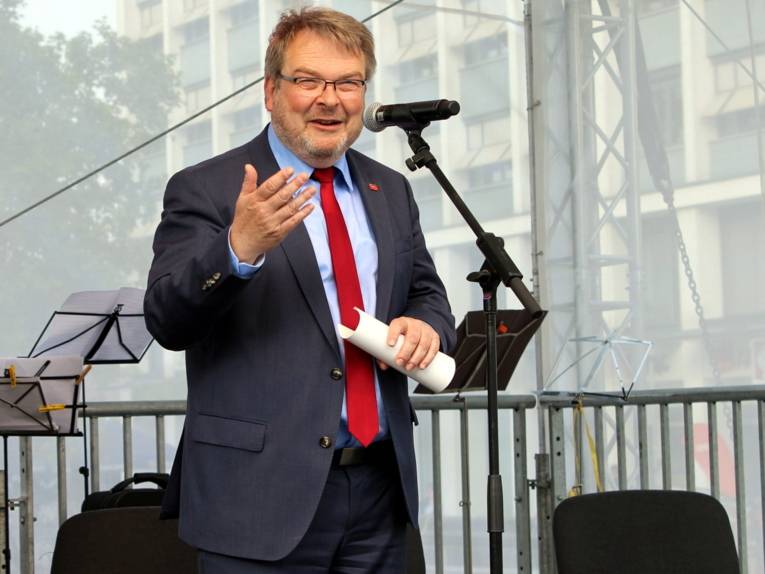 Ein Mann steht auf der Bühne, spricht in ein Mikrofon und gestikuliert mit seiner Hand. In der anderen Hand hält er einen Stapel zusammengerollter Papiere. 