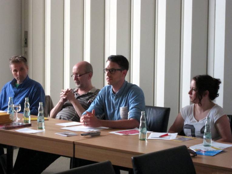 Vier Personen sitzen nebeneinander an Tischen. Die zweite Person vom rechts spricht.