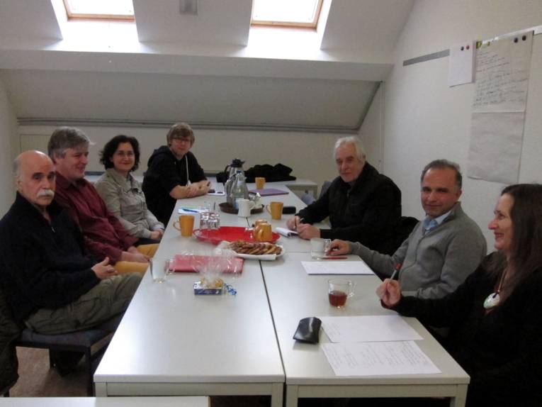 Sieben Personen sitzen um rechteckig aufgestellte Tische. Auf den Tischen sind Süßigkeiten und Kannen und Becher.