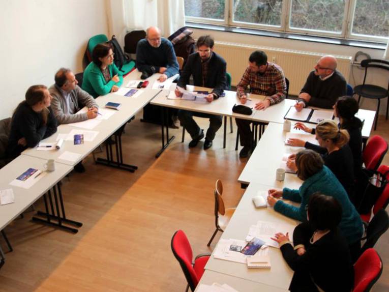 Auf dem von schräg oben aufgenommenen Bild sieht man elf Personen, die um U-förmig aufgestellte Tische sitzen und zu einer Person schauen, die auf einen Zettel in der Hand schaut.