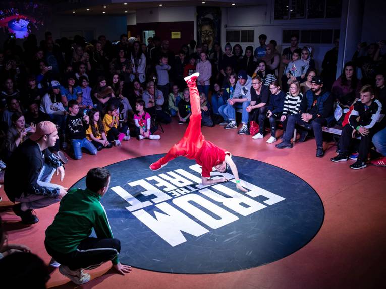 Ein Jugendlicher stützt sich mit Ellenbogen und Hand auf der Tanzfläche ab und wirbelt mit den Beinen durch die Luft; im Umkreis darum feuern ihn die meist jungen Zuschauerinnen und Zuschauer an.
