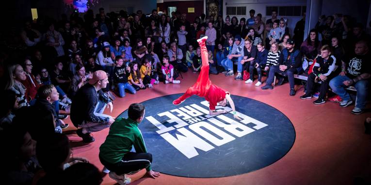 Ein Jugendlicher stützt sich mit Ellenbogen und Hand auf der Tanzfläche ab und wirbelt mit den Beinen durch die Luft; im Umkreis darum feuern ihn die meist jungen Zuschauerinnen und Zuschauer an.