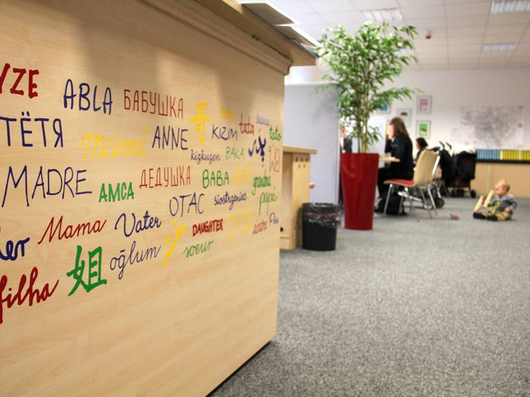 Ein Empfangstresen im Familienservicebüro, auf dem Verwandtschaftsbezeichnungen in unterschiedlichen Sprachen zu sehen sind. Im Hintergrund eine Beratungssituation, bei der ein Kleinkind auf dem Fußboden spielt.