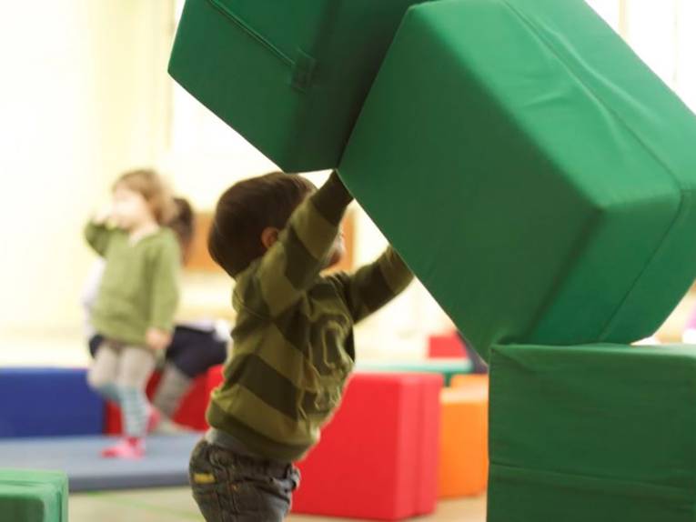 Ein Junge spielt mit übergroßen Schaumstoffwürfeln, im Hintergrund sind verschwommen zwei weitere Kinder zu sehen.
