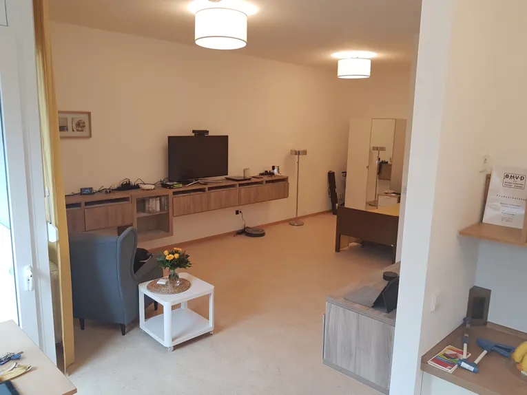 Zimmer mit Sessel, Rollcontainer und an der Wand festgeschraubten Sideboards, auf denen zahlreiche elektronische Geräte zur alltäglichen Unterstützung liegen.