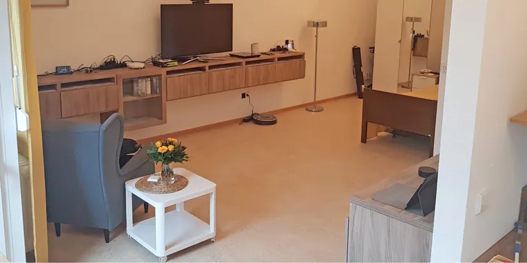 Zimmer mit Sessel, Rollcontainer und an der Wand festgeschraubten Sideboards, auf denen zahlreiche elektronische Geräte zur alltäglichen Unterstützung liegen.