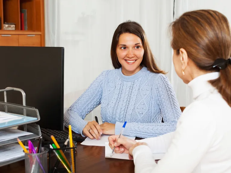 Beratungssituation in einem Büro, bei der sich zwei Frauen mit Stift und Zettel an einem Schreibtisch besprechen.