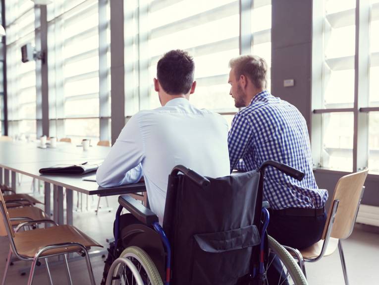 Zwei junge Männer sitzen nebeneinander an einem Besprechungstisch und unterhalten sich; einer davon mit Rollstuhl.