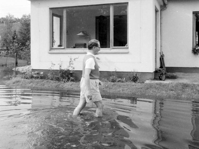 Ein Mann in kurzen Hosen watet bis zu den Waden in Wasser durch seinen Vorgarten.