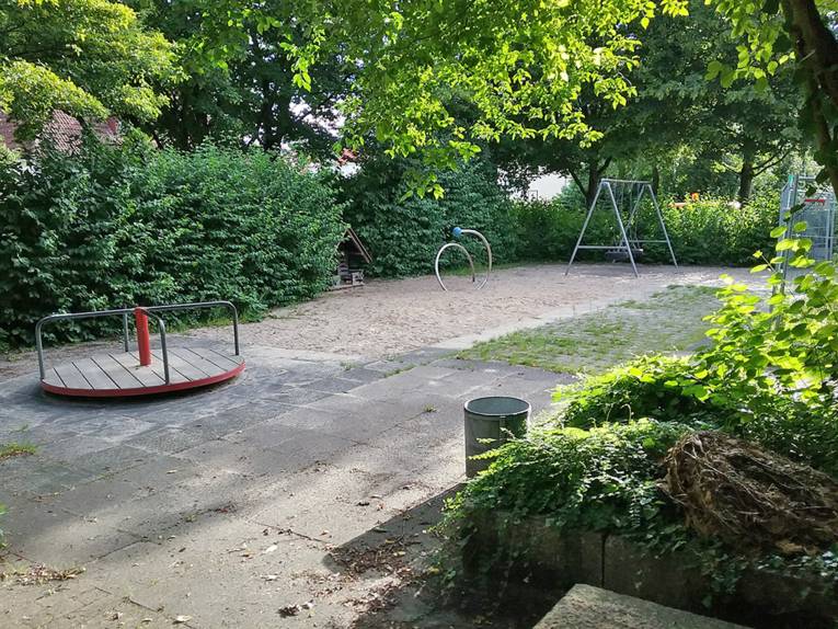 Mehrere Spielgeräte auf einem Spielplatz, der von grünen Büschen umgeben ist