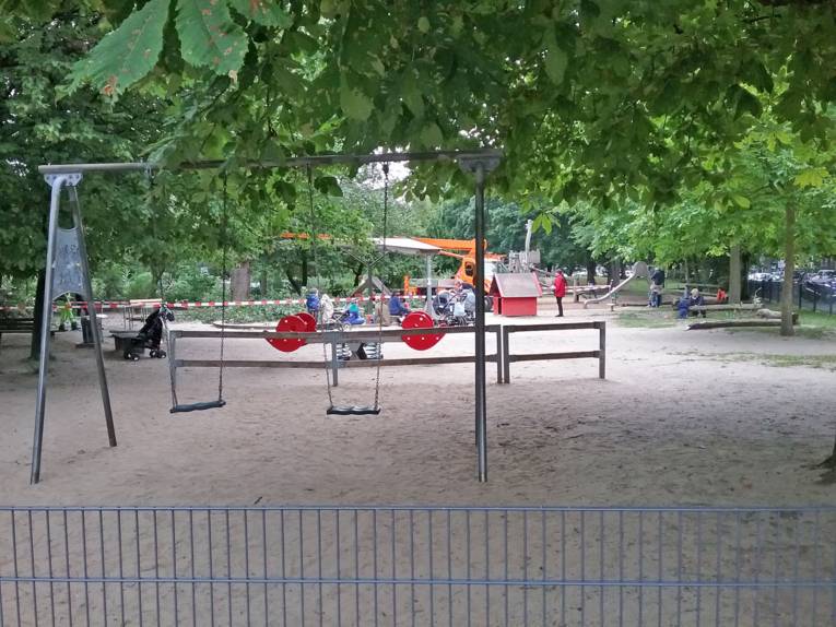 Der Spielplatz mit Spielgeräten wie Schaukel, Wippe und Rutsche befindet sich unter Kastanienbäumen. Kinder spielen dort unter Aufsicht der Erziehungsberechtigten.