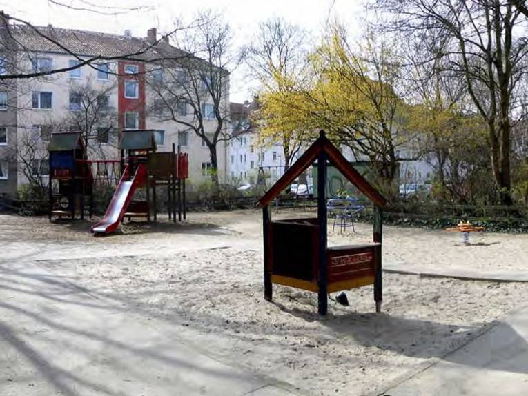 Verschiedene Spielgeräte aus Holz und Metall auf einem mit Sand befüllten Spielplatz, umrahmt von Wohnhäusern