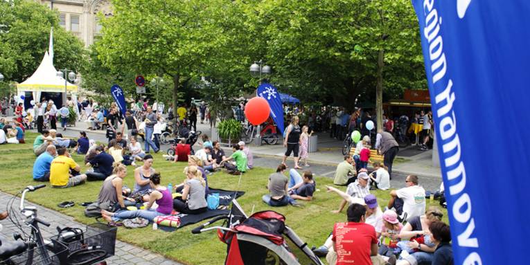Ein Teil der Georgstraße, die mit Rollrasen bedeckt ist, auf der mehrere Dutzend Menschen sitzen und sich unterhalten oder picknicken